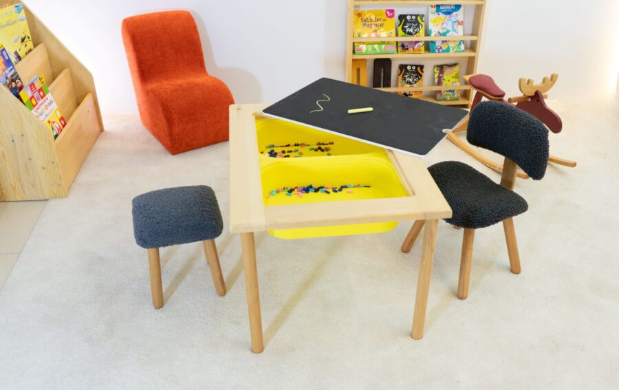 Ahşap çerçeveli Montessori duyusal aktivite masası, beyaz plastik kaplar, kara tahta yüzeyi ve teddy kumaş kaplı sandalyesiyle birlikte kitaplık ve oyuncaklarla dekore edilmiş çocuk odasında yer alıyor.